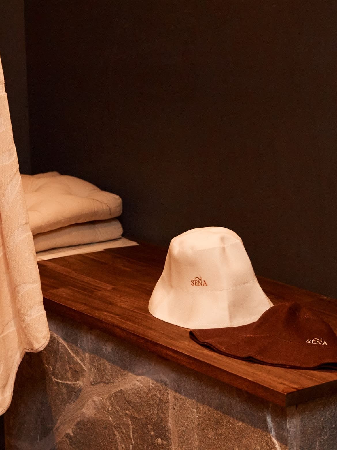A SENA sauna hat and folded towels resting on a timber bench in a warmly lit sauna