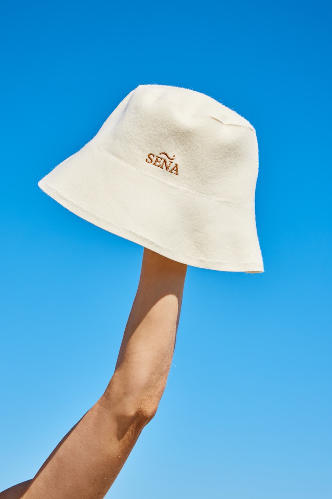 A cream SENA sauna hat held up against a clear blue sky