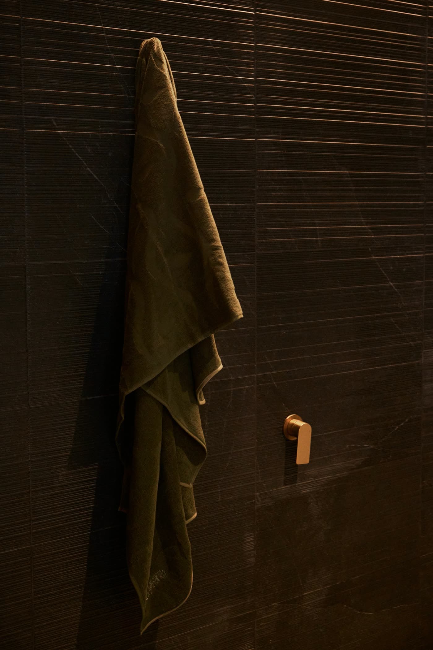 An olive SENA towel hanging from a brass hook on a dark stone wall